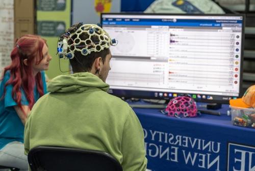 A guest at a health fair wears a brain wave helmet while a neuroscience student monitors the data showing up on a large monitor