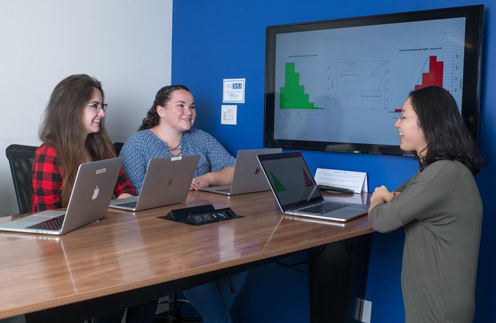 A group of data science students sit at a table with their laptops and graphs on a large monitor